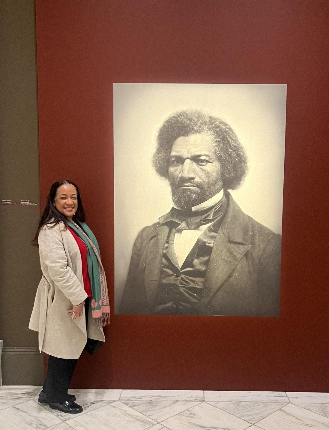 Dr. Carolyn Haliburton Carter standing beside a large gallery reproduction of the well-known daguerreotype portrait of Frederick Douglass.