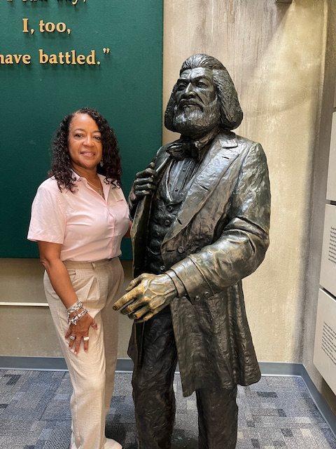 Dr. Carolyn Haliburton Carter standing beside a life-size bronze statue of Frederick Douglass, below a wall bearing the words “I, too, have battled”.