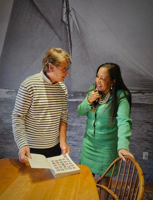 Dr. Carolyn Haliburton Carter and Paul Elias standing at a table, turning the pages of a large album together, in a room hung with a photograph of a sailing ship.