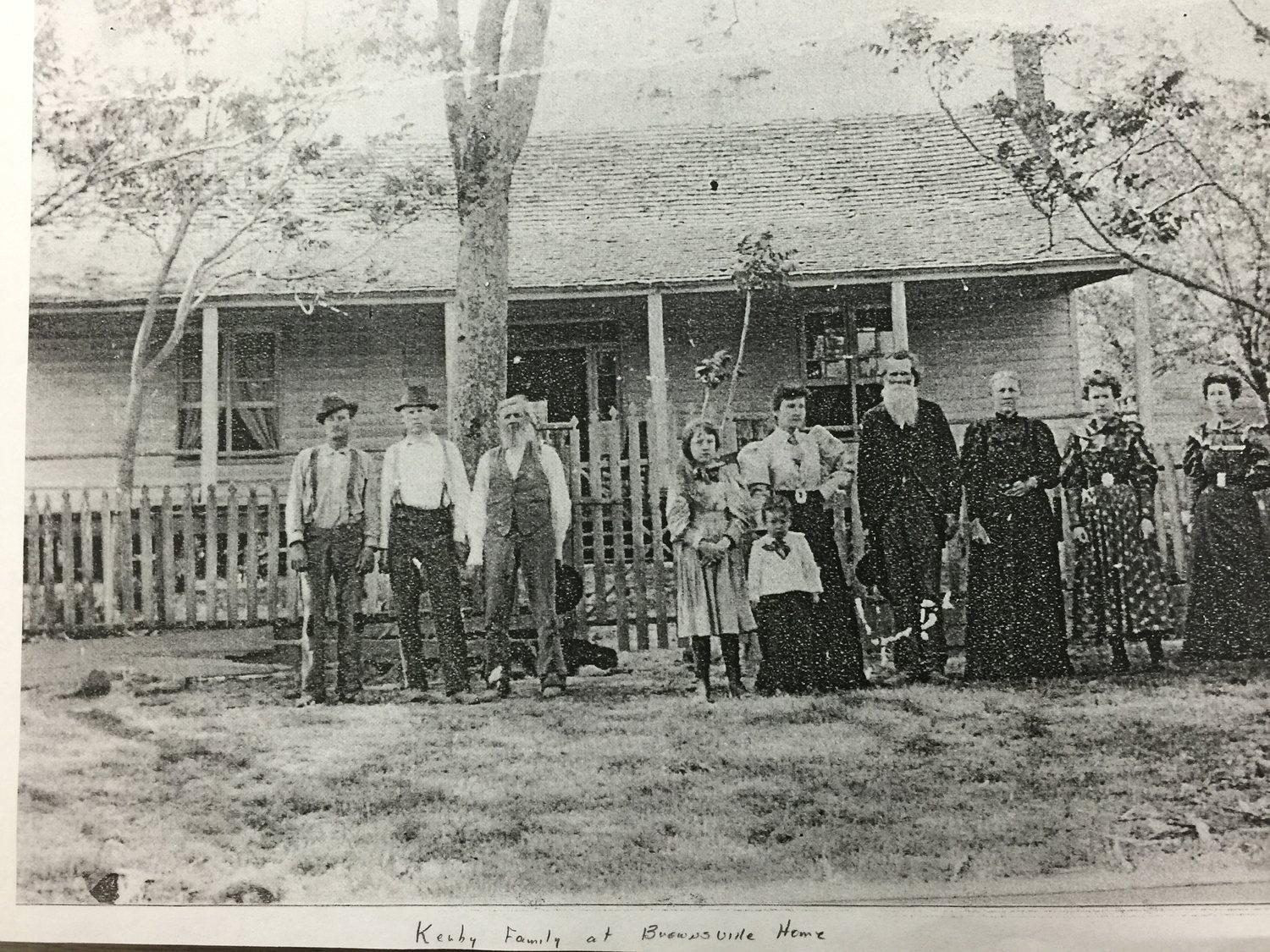 A copy photograph of a late nineteenth-century family group standing outside a timber house behind a picket fence, captioned in handwriting “Kerby Family at Brownsville Home”.
