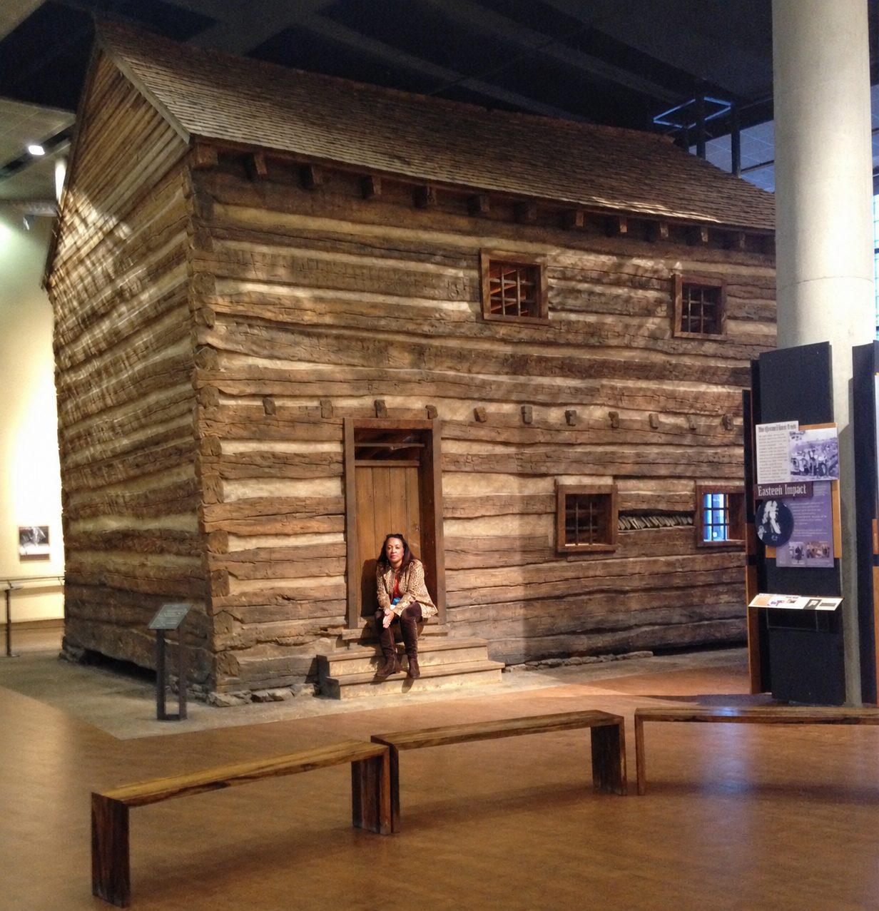 Dr. Carolyn Haliburton Carter sitting on the steps of the two-storey log slave pen installed inside the National Underground Railroad Freedom Center in Cincinnati, with museum benches and an interpretive panel nearby.
