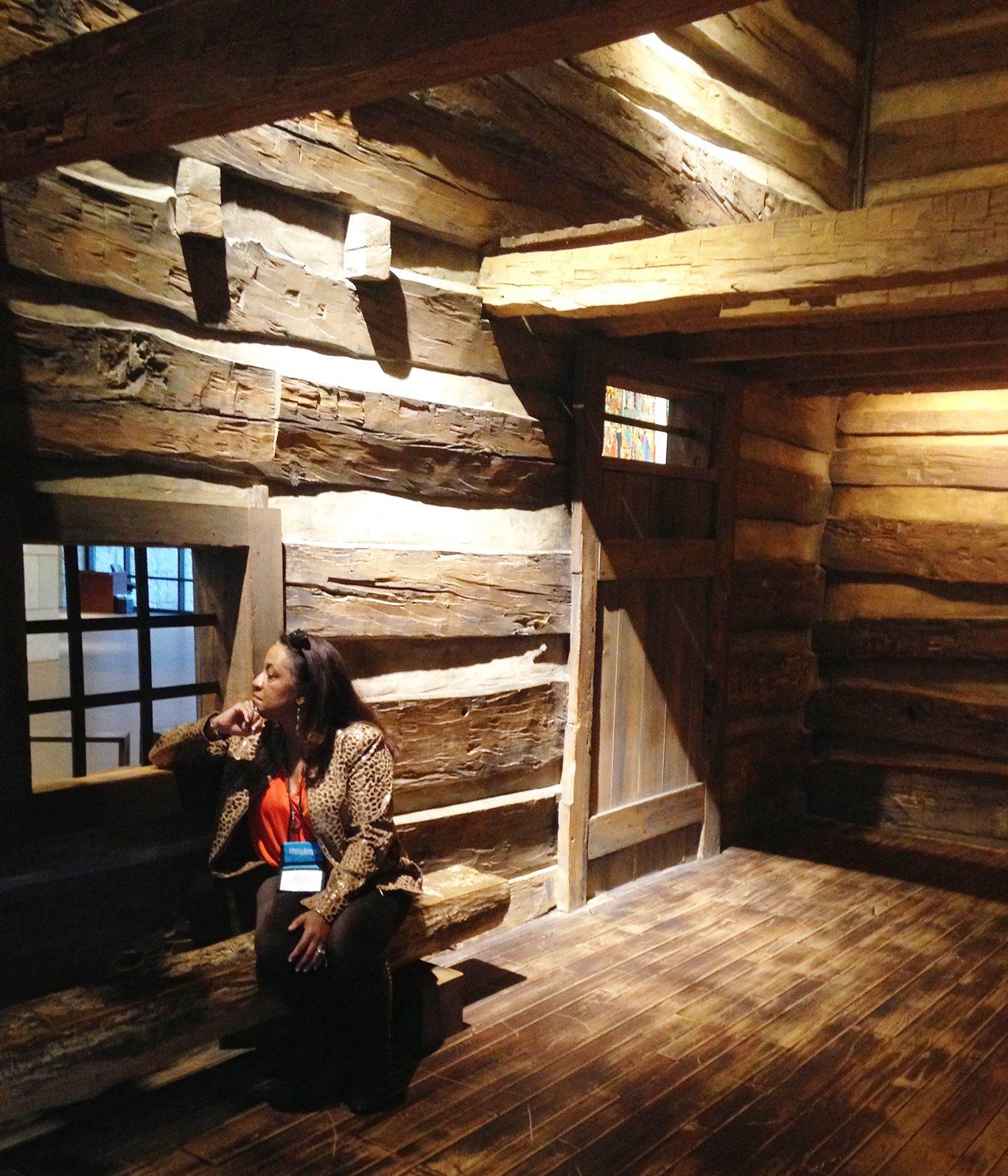 Dr. Carolyn Haliburton Carter sitting alone on a low log ledge inside the slave pen, chin resting on one hand, surrounded by hand-hewn log walls, with a small window beside her, a plank door to the right and a loft overhead.